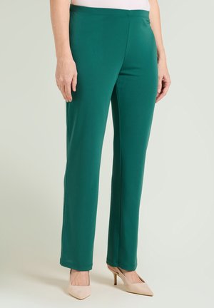Femme portant un pantalon vert à jambes droites et des chaussures beiges à talons hauts, debout devant un fond clair.