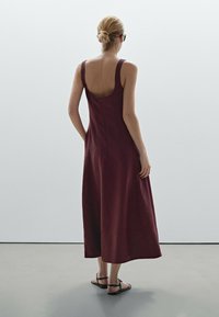 Burgundy-linjan midi-mekko leveillä olkaimilla, syvällä selällä ja huomaamattomalla vetoketjulla. Yhdistetty mustien matalapohjaisten sandaalien kanssa neutraalilla taustalla.