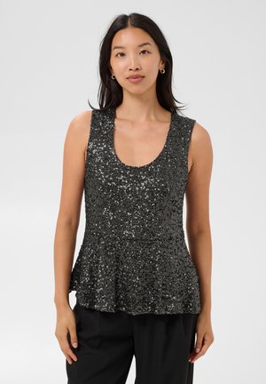 Femme portant un haut noir sans manches à sequins avec un col dégagé et un pantalon noir, debout devant un fond clair uni.