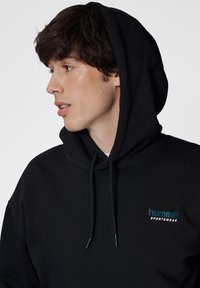 Zwarte hoodie gemaakt van zachte stof met een voorzak in kangaroo-stijl, voorzien van een turquoise "hummel" logo op de linkerborst. Verstelbare trekkoordkap.