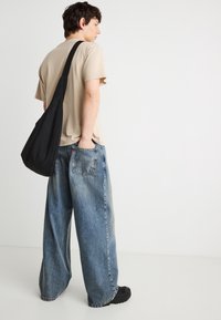 Νεαρός άντρας που στέκεται πλάγια φορώντας μπεζ μπλουζάκι, φαρδιά μπλε τζιν, μαύρα παπούτσια και μαύρη τσάντα χιαστί σε λευκό φόντο.