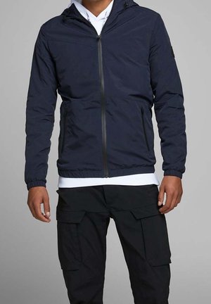 Homme portant une veste à capuche zippée bleu marine sur une chemise blanche avec un pantalon cargo noir, debout devant un fond gris uni.