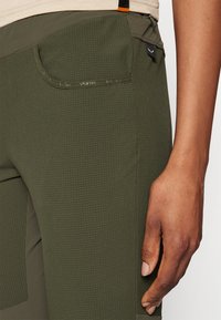 Olive green pants featuring a textured pattern, side pocket, and contrasting dark fabric accents at the hem and waist.