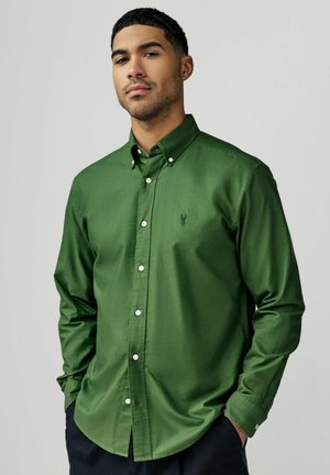Chemise verte à boutons, en tissu lisse, avec un col pointu, des boutons sur le devant et un petit logo sur la poitrine gauche.
