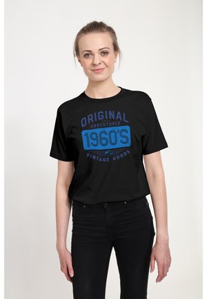Kvinde med lyst hår iført en sort t-shirt med teksten "Original and Unrestored 1960's Vintage Goods" og sorte bukser, stående foran en hvid baggrund.