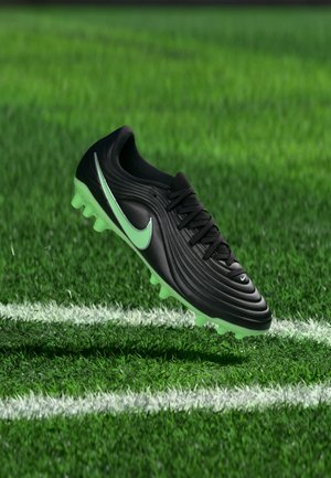 Μαύρο και πράσινο παπούτσι ποδοσφαίρου Nike αιωρείται πάνω από μια λευκή γραμμή πάνω σε πράσινο χλοοτάπητα.