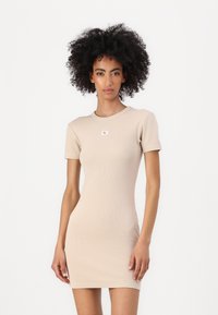 Beige ribbed dress with short sleeves, fitted silhouette, crew neckline, and a small branded tag on the chest. Soft, stretchy fabric.