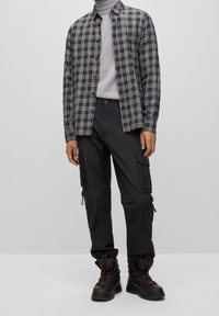 Schwarzes kariertes Flanellhemd über einem grauen Rollkragenpullover, kombiniert mit schwarzen Cargo-Hosen mit Reißverschlüssen und Taschen sowie schwarzen Wanderschuhen.