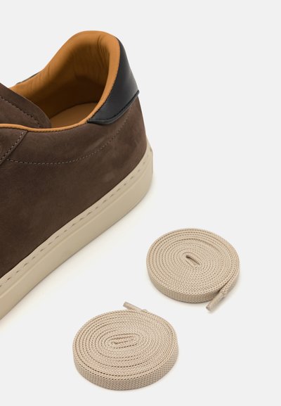 Sneaker in suede marrone con un accento in pelle nera sul tallone, suola in gomma beige e due lacci beige arrotolati accanto alla scarpa.