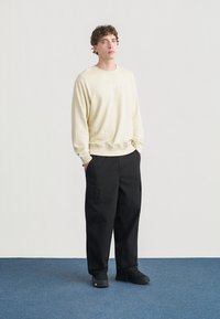 Ανοιχτό μπεζ φούτερ με στρογγυλή λαιμόκοψη και ριμπ μανσέτες, σε συνδυασμό με φαρδιά μαύρη παντελόνια cargo με πλαϊνές τσέπες και ραφές.