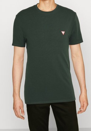 Homme portant un t-shirt à manches courtes vert foncé avec un petit logo triangulaire rouge et blanc sur la poitrine gauche, associé à un pantalon noir.
