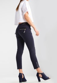 Vaqueros azul marino ajustados con cremalleras laterales, combinados con una blusa corta blanca y tacones destalonados azul marino. Tejido liso y elástico, diseño estilizado.