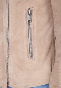 Beige suède jasje met een zijritszak, zilveren hardware en zichtbare stiksels, wat een soepele textuur benadrukt.