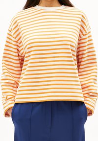 Gestreiftes Langarmshirt aus orange-weißem Baumwollstoff, mit Rundhalsausschnitt, lockerer Passform und sichtbaren Nähdetails. Kombiniert mit marineblauen Hosen.