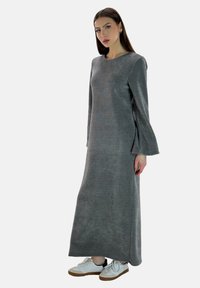 Vestido maxi gris de manga larga en una tela suave, con una silueta relajada, aberturas laterales y un escote redondo. Combinado con zapatillas blancas.