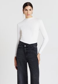 Frau in weißem, geripptem Langarmoberteil und hochgeschnittenen schwarzen Jeans, die vor einem schlichten weißen Hintergrund steht.