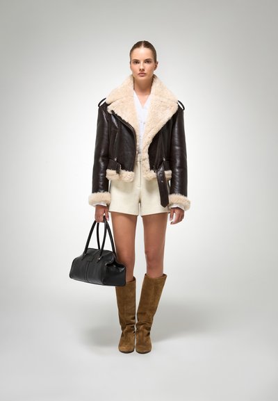 Bruine leren cropped jacket met een crèmekleurige nepbontkraag, witte top, beige shorts, kniehoge bruine suède laarzen en een zwarte handtas.
