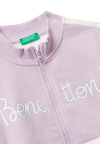 Jasnopurpurowy sweter zapinany na zamek z prążkowanym kołnierzem i napisem "Benetton" wyszytym na białym tle na klatce piersiowej, z zieloną metką "United Colors of Benetton".