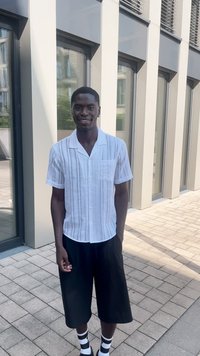 Camisa branca de manga curta com botões e riscas verticais, combinada com calções pretos folgados e meias às riscas. De pé sobre uma superfície pavimentada.