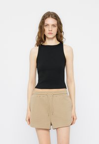 Γυναίκα με μακριά κυματιστά μαλλιά που φοράει ένα μαύρο αμάνικο crop top και μπεζ σορτς με κορδόνι, στέκεται μπροστά σε ένα απλό λευκό φόντο.