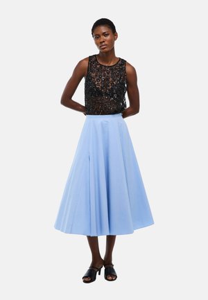 Femme portant un haut noir sans manches orné de perles et une jupe midi bleue taille haute, debout les mains dans le dos sur fond blanc.