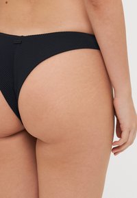 Juodos juostos bikini kelnaitės su minimalizmo dizainu, plona juosta ir nepastebimais kraštais patogumui. Matoma subtili tekstūra ir pritaikymo detalės.