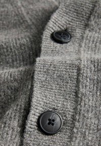 Cárdigan gris de punto con textura acanalada, dos grandes botones negros y un material suave similar a la lana. Primer plano del botón y el detalle de la tela.