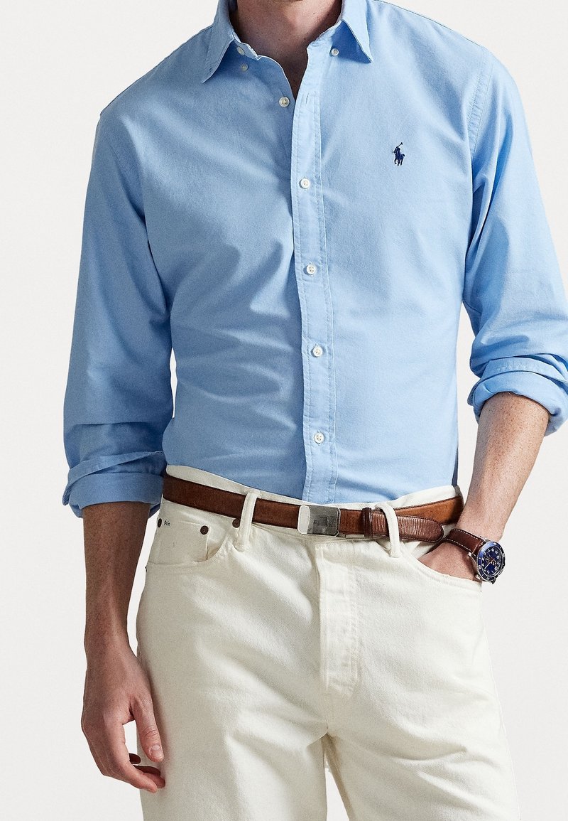 Homme portant une chemise bleu clair boutonnée avec les manches retroussées, un pantalon beige, une ceinture en cuir marron et une montre foncée au poignet gauche.