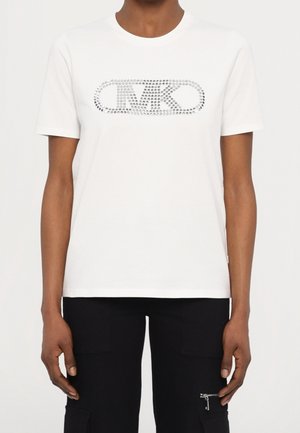 Camisa blanca de manga corta con logo ovalado adornado con piedras brillantes en el pecho, llevada por una persona de piel oscura y pantalones negros con detalle de cremallera.