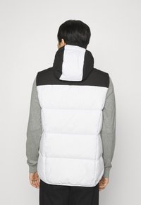 Putlinis liemenė su juodai baltu spalvų blokų dizainu. Yra aukštas apykaklė, nuimama kapa ir tekstūruota medžiaga.