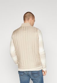 Gilet matelassé beige avec un col côtelé et un design sans manches, associé à une chemise à manches longues légère et texturée, ainsi qu'à un jean en denim.