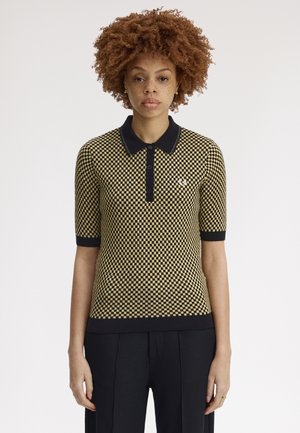 Femme aux cheveux bouclés portant un polo à manches courtes à carreaux jaunes et noirs et un pantalon noir, debout devant un fond uni.