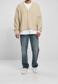 Cardigan de punto beige con cuello en V y cierre de botones, usado sobre una camisa blanca, combinado con jeans azules y zapatillas blancas.