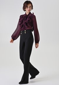 Bordeauxrode blouse met ruches en transparante stof, gecombineerd met zwart, hooggetailleerde broek met gouden knopen en een uitlopende zoom.