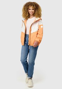 Orange och vit zip-jacka med huva, justerbara dragsnören och elastiska ärmslut, i kombination med blå jeans och vita sneakers.