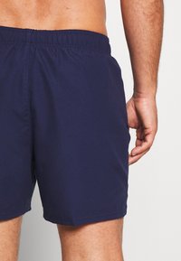 Shorts de bain bleu marine foncé avec une taille élastique, texture lisse et sans motifs visibles, montrant une main positionnée sur le côté.