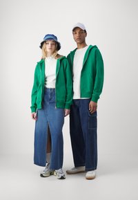 Des sweats à capuche zippés verts portés sur un haut blanc ; associés à une jupe en denim et un jean ample. Les accessoires incluent un chapeau de seau bleu et une casquette blanche.