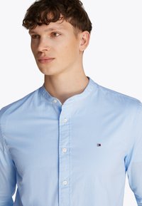 Ung mann med kort brunt hår iført en lyseblå skjorte med mandarin-krage, hvite knapper og en liten logo på brystet.