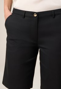 Shorts noirs en tissu lisse, dotés d'une poche avant, d'une fermeture à bouton et de coutures soignées le long de l'ourlet. Design simple et ajusté.