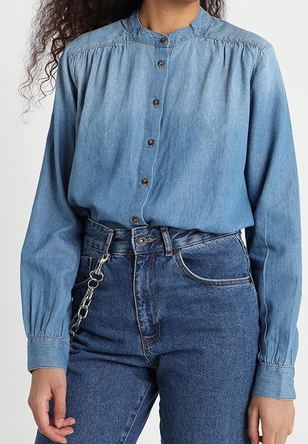 Personne portant une chemise en jean bleu clair boutonnée, rentrée dans un jean bleu foncé taille haute, avec une chaîne argentée suspendue à la taille.