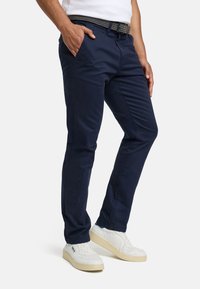 Pantaloni chino din bumbac bleumarin cu buzunare, croială conică, având o curea țesută cu model și pantofi sport albi cu tălpi bej.