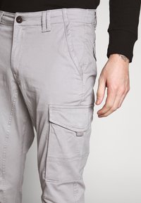 Man in een lichtgrijze cargobroek met zijzakken en een zwarte long sleeve shirt zichtbaar bij de pols.