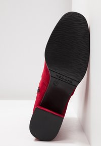Chaussure en daim rouge avec semelle texturée noire et talon bloc, inclinée vers le haut montrant la marque "Tamaris" et la taille 37 sur la semelle.