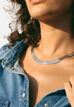 Silver metal necklace with a curved, flat design. Worn over a denim shirt, featuring a smooth texture and subtle shine.