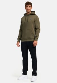 Mann steht und trägt eine olivgrüne Kapuzenjacke, schwarze Hose und weiße Turnschuhe vor einem schlichten weißen Hintergrund.