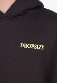 Sweat-shirt noir avec le texte "DROPSIZE" en couleur crème imprimé sur la poitrine, montrant une partie de la capuche et du col.