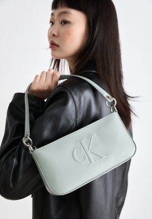 Ung kvinde med glat mørkt hår bærer en sort læderjakke og har en lyseblå Calvin Klein skuldertaske med prægede logo.