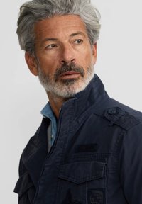 Chaqueta de estilo militar azul marino con cuello alto, múltiples bolsillos, botones y tejido texturizado. Llevada sobre una camisa de azul claro.