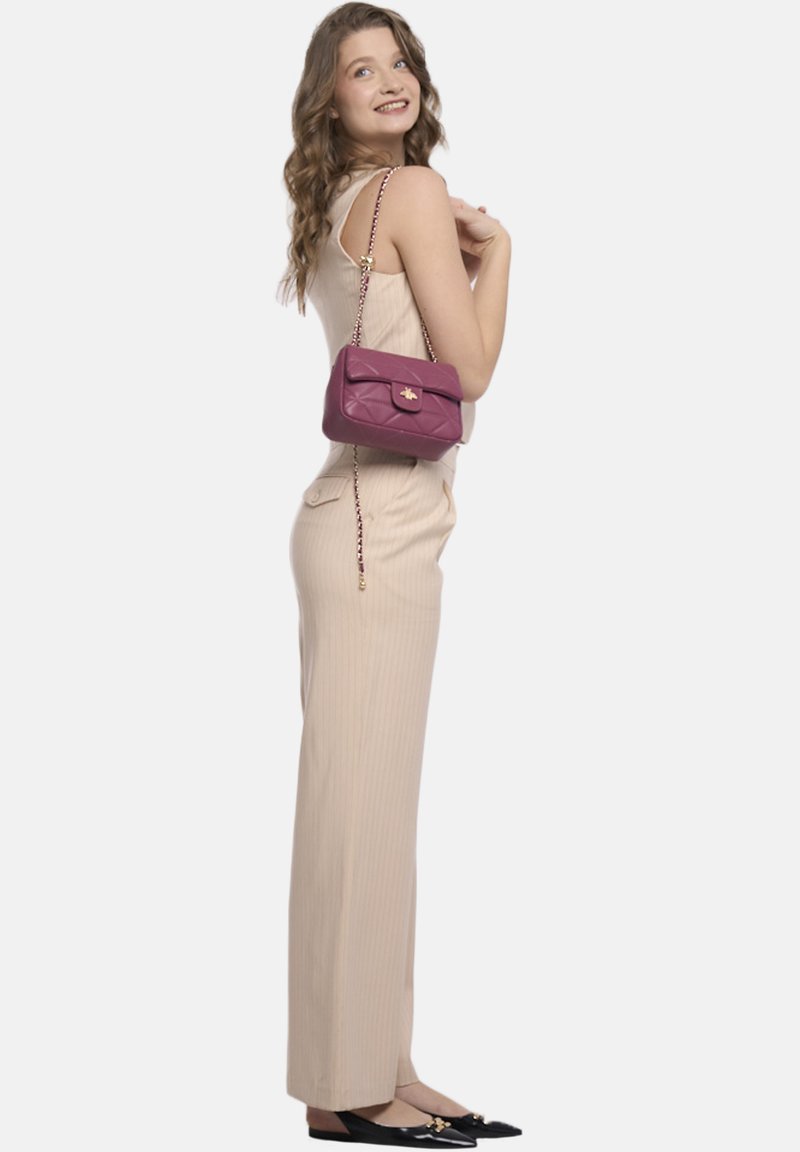 Jumpsuit a coste color beige chiaro, abbinato a una piccola borsa a tracolla bordeaux trapuntata con chiusura dorata e tracolla a catena. Ballerine nere completano il look.