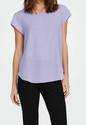 Kobieta ubrana w lawendową bluzkę z krótkim rękawem i okrągłym dekoltem oraz czarne spodnie, stojąca na biały tle.